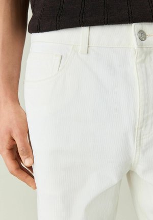 Personne portant un pantalon blanc texturé et un haut noir côtelé, la main gauche reposant près de la poche avant du pantalon.