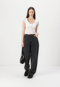 Witte kanten crop top met een knoopdetail, gecombineerd met losse zwarte jeans met witte spikkels. Model houdt een zwarte schoudertas vast.