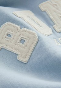 Világoskék szövet, textúrált, hímzett fehér "89" számmal, négyzet alakú kivágásokkal és varrt díszítésekkel, amelyek részletes kézműves munkát mutatnak.