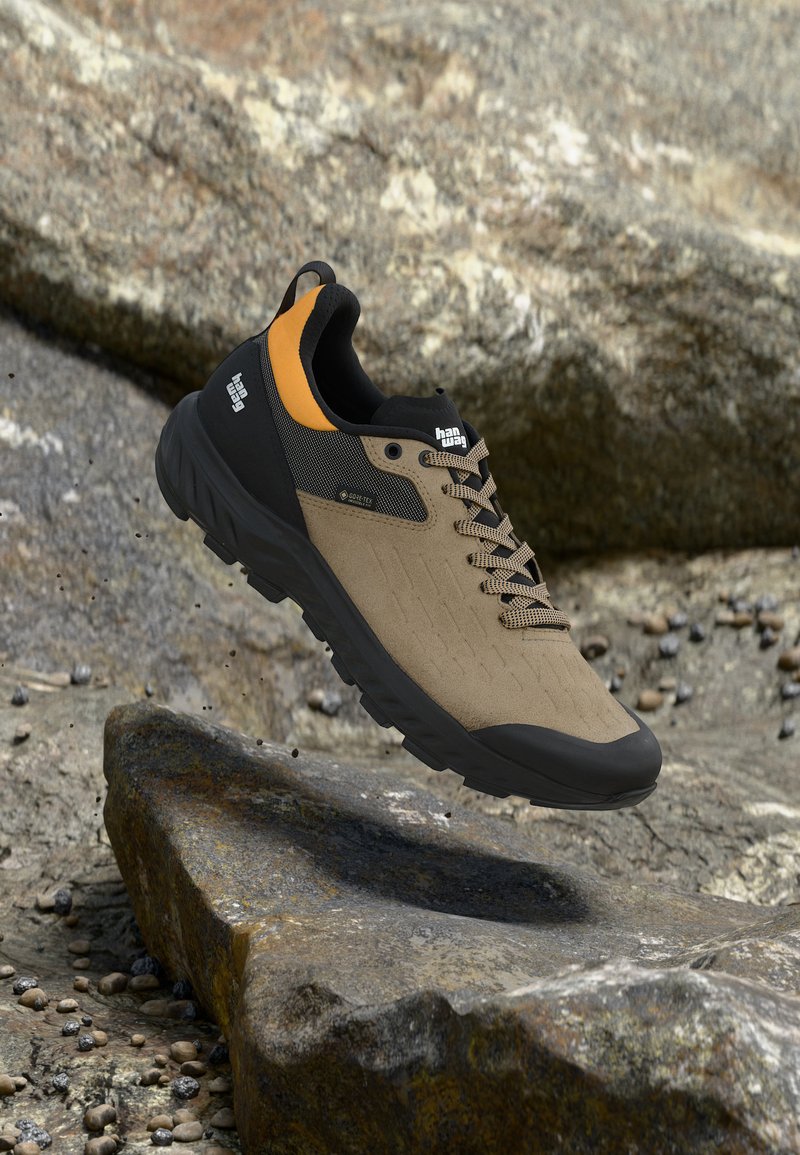 Μπεζ και μαύρο πεζοπορικό παπούτσι με πορτοκαλί λεπτομέρειες, χοντρή σόλα και κορδόνια, αιωρούμενο πάνω από πετρώδες υπαίθριο έδαφος.