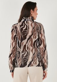 Blusa trasparente con colletto arricciato e maniche lunghe, caratterizzata da un motivo ondulato marrone, beige e bianco. Chiusura con bottoni ai polsini.