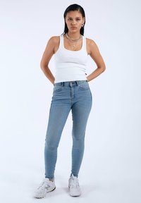 Top de tirantes blanco acanalado combinado con jeans ajustados de color azul claro. Se lleva con zapatillas blancas que tienen una suela texturizada y detalles metálicos.