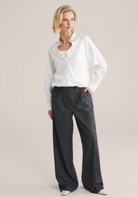 Camicia bianca con maniche a manica lunga, abbinata a pantaloni larghi e scuri dalla texture morbida, e scarpe bianche con punta a forma di freccia.