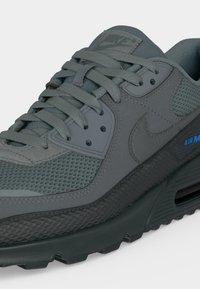 Γκρι αθλητικό παπούτσι Nike Air Max με πάνελ από δίχτυ και δέρμα, υφή στη σόλα, και μπλε λογότυπο «Air Max» στο πλάι.