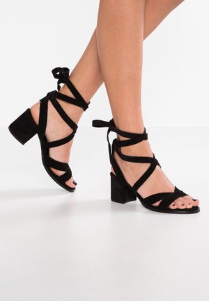 Czarne zamszowe sandały na obcasie mają plecioną konstrukcję z wiązaniami na kostkach. Obcasy mają kształt bloku, co zapewnia stabilność i wsparcie.