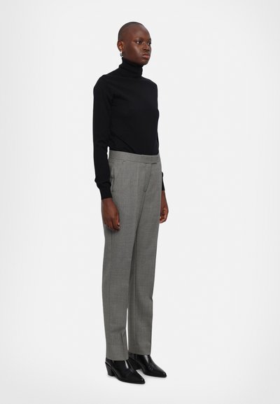 Crna turtleneck pulover, sivi karirani hlače s krojem, i crne čizme s potpeticama do gležnjeva. Model stoji uspravno protiv svijetle pozadine.