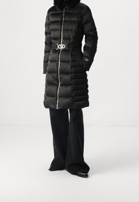 Abrigo largo acolchado negro con cremallera plateada y cinturón, llevado sobre pantalones negros de pierna ancha y zapatos negros puntiagudos sobre un fondo liso.