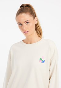 Sweatshirt beige clair avec un col rond, affichant un design de logo multicolore sur la poitrine. Texture lisse, coupe décontractée et manches longues.