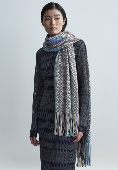 Geweven sjaal met een multicolor zigzagpatroon en franjes aan de uiteinden. Gemaakt van lichtgewicht materiaal, tentoongesteld over een donker gebreide outfit met patroon.