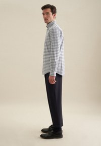Camicia a quadri azzurro chiaro e bianco con colletto, abbinata a pantaloni scuri e scarpe nere. Il modello è in posizione laterale, con le mani rilassate.