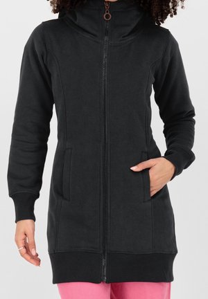 Femme portant un long sweat à capuche noir zippé avec des poches, la main droite dans une poche et la main gauche détendue le long du corps.