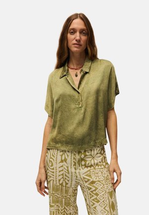 Mujer con cabello largo y castaño que viste una camisa suelta de manga corta color verde oliva y pantalones beige y verdes con estampado sobre un fondo liso.