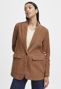 Brown corduroy blazer with two front pockets, notch lapels, and a single button closure. Paired with a light beige V-neck sweater.