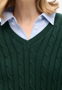 Woman wearing a green cable-knit V-neck sweater over a blue and white striped collared shirt, shown from shoulders to neck.
