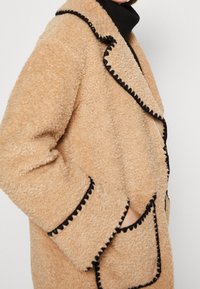 Beżowy płaszcz pluszowy z kontrastową czarną lamówką, z falistymi krawędziami, dwoma przednimi kieszeniami i miękkim, teksturowanym materiałem.