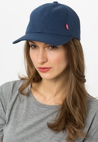Marineblå baseball cap med strukturelt design og buet skygge, med et lite rødt logomerke på siden. Laget av bomull.