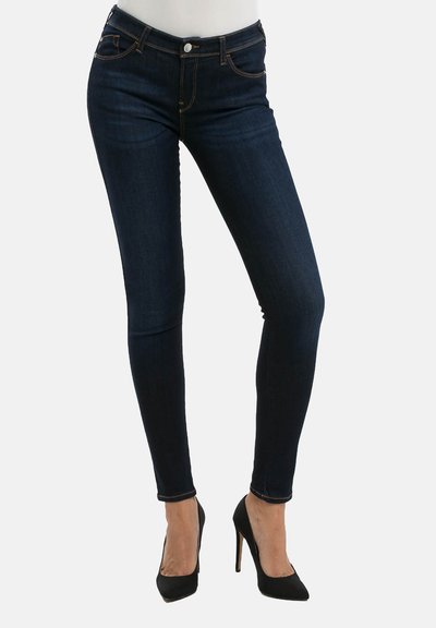 Jeans skinny azul oscuro de mezclilla, con una cintura alta, bolsillos frontales y costuras naranjas contrastantes. Combinados con tacones negros.