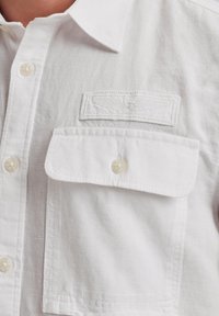 Camisa de lino blanca con cuello abotonado, presenta un bolsillo parche y un logo bordado. Textura suave con botones de perlas.