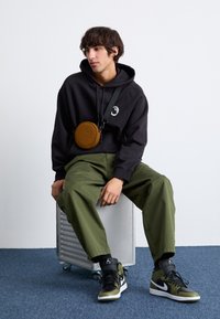 Black hooded sweatshirt with a logo, olive green loose-fit pants, and black/olive high-top sneakers. Circular brown bag with a strap.
