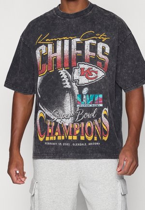 Maglietta nera dei campioni del Super Bowl LVII dei Kansas City Chiefs con grafica di football, indossata da una persona con pantaloncini cargo grigio chiaro.