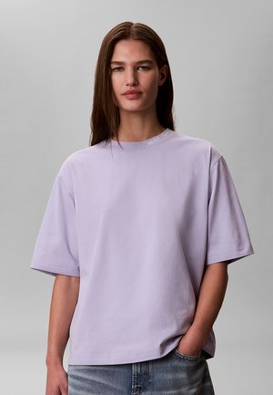 Ung kvinde med langt brunt hår iført en løs, lyse lilla T-shirt og blå jeans, stående mod en ensfarvet grå baggrund.