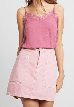 Femme portant un top rose sans manches orné de dentelle, rentré dans une jupe rose clair taille haute avec de grandes poches avant.