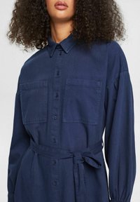 Abito camicia blu navy realizzato in tessuto morbido. Presenta due tasche sul petto, una chiusura con bottoni frontale e una vita legata per una silhouette aderente.