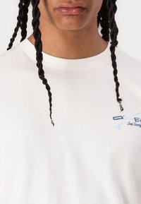 Hvid bomulds T-shirt med et blåt grafisk logo med tekst på venstre side. Rund halsudskæring og korte ærmer. Glat tekstur.