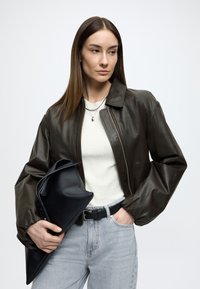 Casaco bomber de couro castanho escuro com gola alta, combinado com uma camisola cinzento claro com nervuras e jeans claros. A segurar uma mala de mão preta.