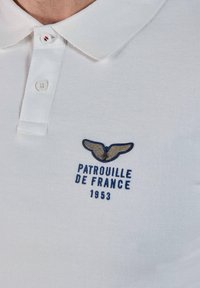 Polo blanc avec col boutonné et logo brodé comprenant des ailes, une étoile et le texte "Patrouille de France 1953" sur la poitrine.