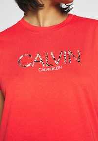 Red cotton t-shirt featuring a shimmered "CALVIN" logo in black and white floral print, with a crew neck and short sleeves.