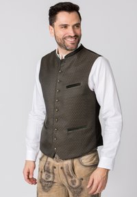 Dunkel gemusterte Weste mit vertikalen grünen Akzenten und dekorativen Knöpfen. Über einem weißen Hemd getragen, kombiniert mit hellen Hosen.