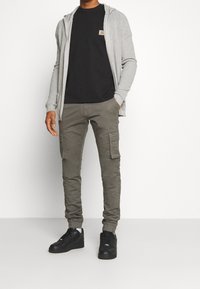 Šedý žebrovaný dlouhý kardigan přes černé tričko, kombinovaný s úzkými olivovými cargo kalhotami a černými teniskami, které mají boční kapsy.