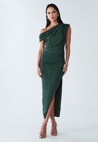 Robe verte à épaules dénudées avec texture froncée, fente jusqu'au genou et longueur au sol. Associée à des talons dorés à brides. Tissu lisse.