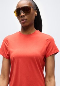 Γυναίκα που φορά ένα φωτεινό κόκκινο κοντομάνικο πουκάμισο και μεγάλα αθλητικά γυαλιά ηλίου με κεχριμπαρένιο φακό και διάφανη κορνίζα, στέκεται μπροστά σε ένα απλό φόντο.