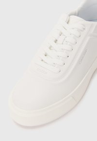 Valged sneakersid sujuvalt sünteetilisest ülemisest, ümar nina, paeltega sulgemine ja külje peal reljeefne brändi logo. Kummist välistald kerge tõusuga.
