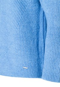 Hellblauer Strickpullover mit geripptem Muster und einem kleinen metallischen Markenschild in der unteren rechten Ecke.