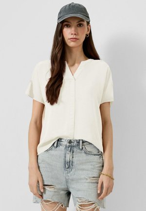 Femme portant un chemisier blanc à manches courtes, un short en jean clair déchiré, une casquette de baseball grise et un bracelet en or, debout sur un fond uni.