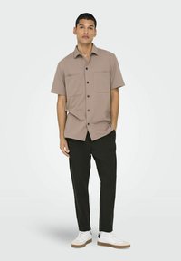 Camisa de botones de manga corta en color beige con dos bolsillos en el pecho, combinada con pantalones negros y zapatillas blancas. Diseño simple y forma ajustada.