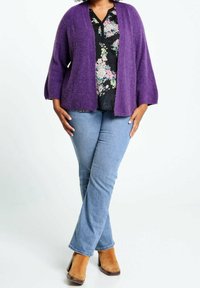 Cardigan viola in un tessuto morbido e strutturato, abbinato a una blusa nera floreale, jeans azzurri e stivali con tacco color cuoio.