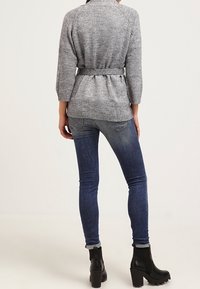 Šedý pletený kardigan s páskem v pase, tříčtvrtečními rukávy a střední délkou, kombinovaný s tmavě modrými džínami a černými kotníkovými botami.