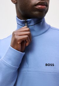 Άνδρας που φοράει ανοιχτό μπλε φούτερ BOSS με φερμουάρ μέχρι το στήθος, τραβάει το φερμουάρ κοντά στον γιακά, κοντινή λήψη στο πάνω μέρος του κορμού και τα χείλη.