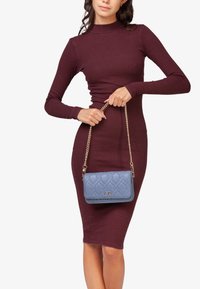 Gequilte lichtblauwe handtas met een gouden kettingriem. Heeft een voorflap met een logo. Draagt door een persoon in een aansluitende bordeauxrode jurk.