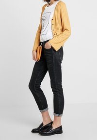 Gul räfflad kardigan över en vit grafisk t-shirt, ihop med uppvikta svarta jeans och svarta loafers. Orange kuvertväska som accent.