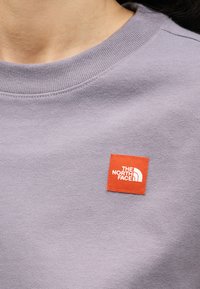 Κοντινό πλάνο ατόμου που φοράει φωτεινό μωβ μπλουζάκι με στρογγυλή λαιμόκοψη και μικρό πορτοκαλί ορθογώνιο ταμπελάκι με το λογότυπο "THE NORTH FACE".