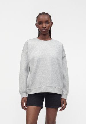 Vrouw met gevlochten haar die een lichtgrijze oversized trui en zwarte spijkershorts draagt, staand tegen een effen witte achtergrond.
