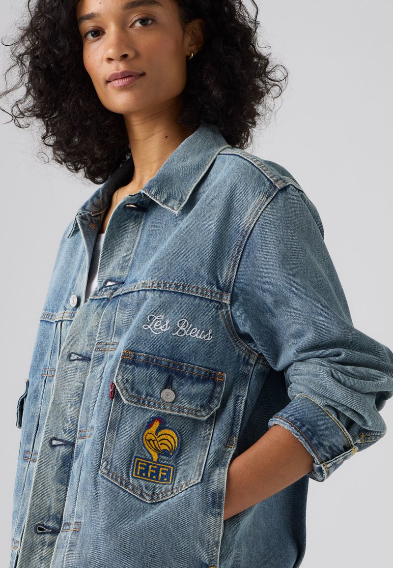 Donna con capelli ricci che indossa una giacca di jeans azzurro chiaro con la scritta "Les Bleus" e una toppa con l'emblema del gallo giallo sulla tasca del petto.