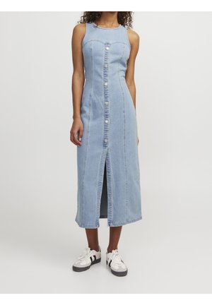 Kvinde iført en ærmeløs, lyseblå denim midi-kjole med knapper foran og en central slids, kombineret med hvide og sorte sneakers.