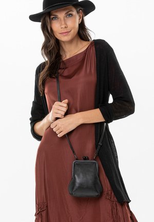 Frau trägt einen schwarzen Hut, eine schwarze Strickjacke und ein rostfarbenes Kleid und hält vor einem weißen Hintergrund eine kleine schwarze Leder-Umhängetasche.