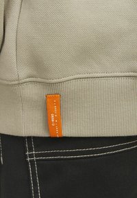 Sweatshirt kaki avec un ourlet côtelé, arborant une étiquette orange bien en vue. Le tissu a une finition texturée, associé à un denim foncé avec des coutures contrastantes.